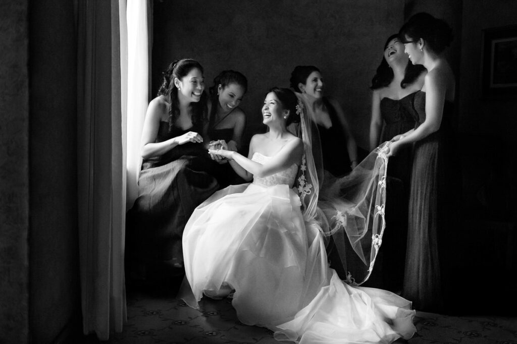 A bride surrounded by bridesmaids laughing together in a dimly lit room, the bride seated and holding onto a veil.