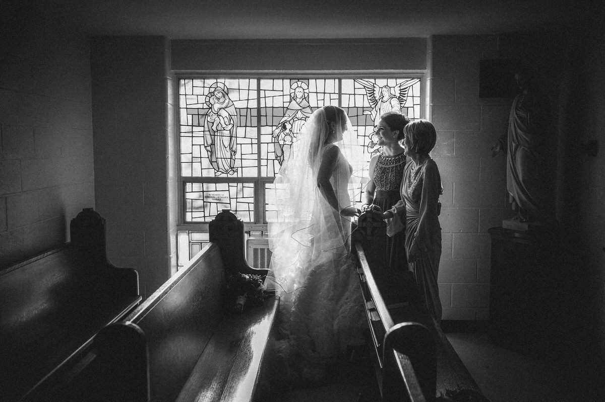 A bride in a white gown stands by stained glass windows, two women adjusting her veil in a serene church setting.