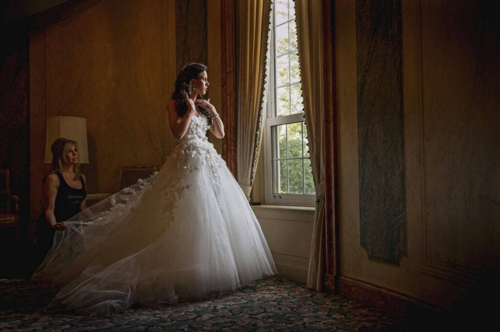 A bride in an elegant white gown standing by a window at Pine Hollow Country Club, with a woman assisting her in the background.