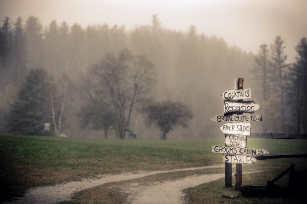 A misty outdoor scene at Riverside Farm featuring a wooden directional signpost listing various wedding venues like "cocktails," "reception," and "stables," next to a muddy path and a wooden