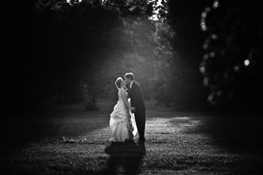 A bride and groom sharing a kiss in a sunlit park, with rays of light creating a halo effect around them during their Please Touch Museum wedding. The scene is captured in black and white photography.