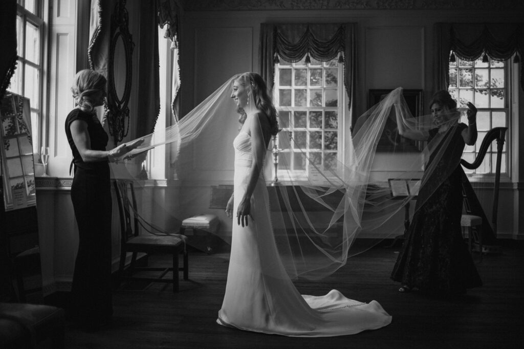 Three women in a room with large windows at Powell House prepare for a wedding; one adjusts a bride's long veil as another watches.