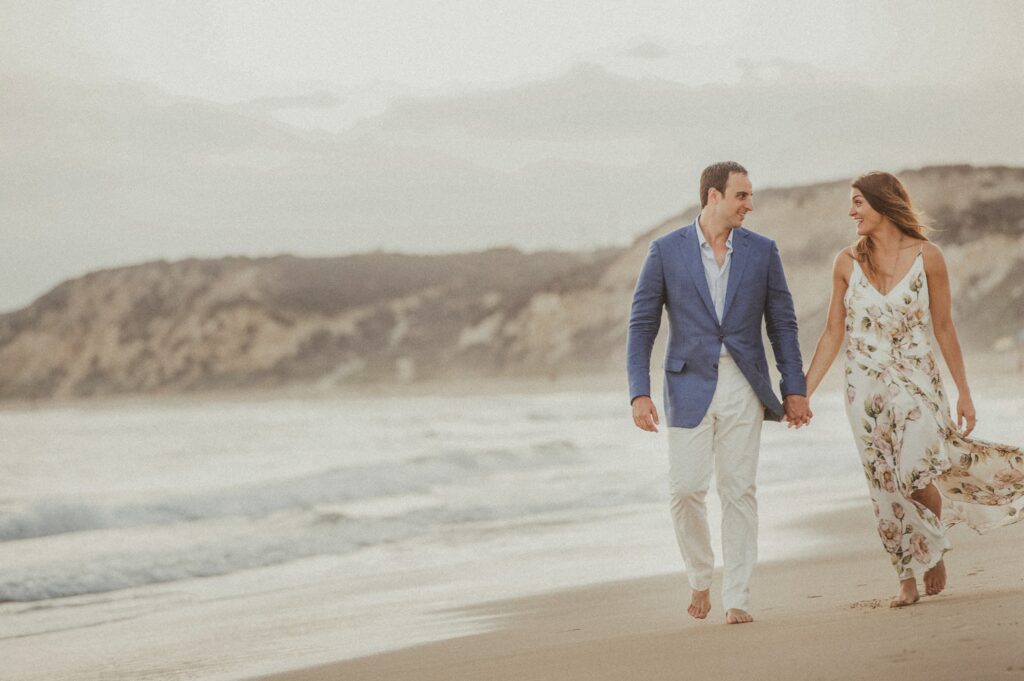A couple holding hands and walking along a sandy beach with cliffs in the background at a Resort at Pelican Hill wedding.