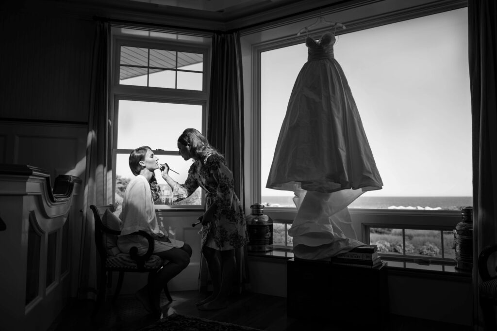 A woman is getting her makeup done by another woman in a room with a view of the ocean at Ocean House in Rhode Island, while a wedding dress hangs by the window.