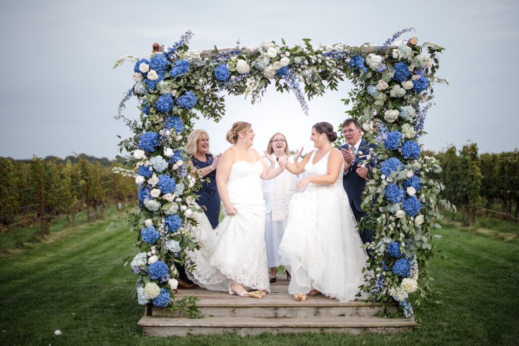 A joyful wedding group, featuring two brides and three guests, laughing under a floral arch at Saltwater Farms Vineyard.