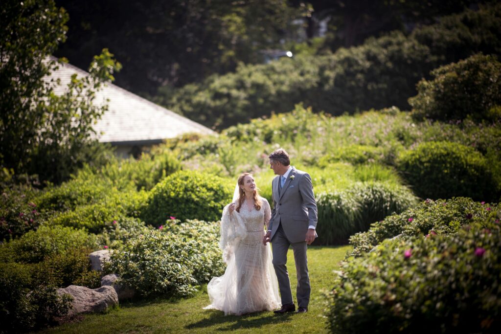 A bride and groom holding hands in an ocean house garden in Rhode Island on a sunny day.