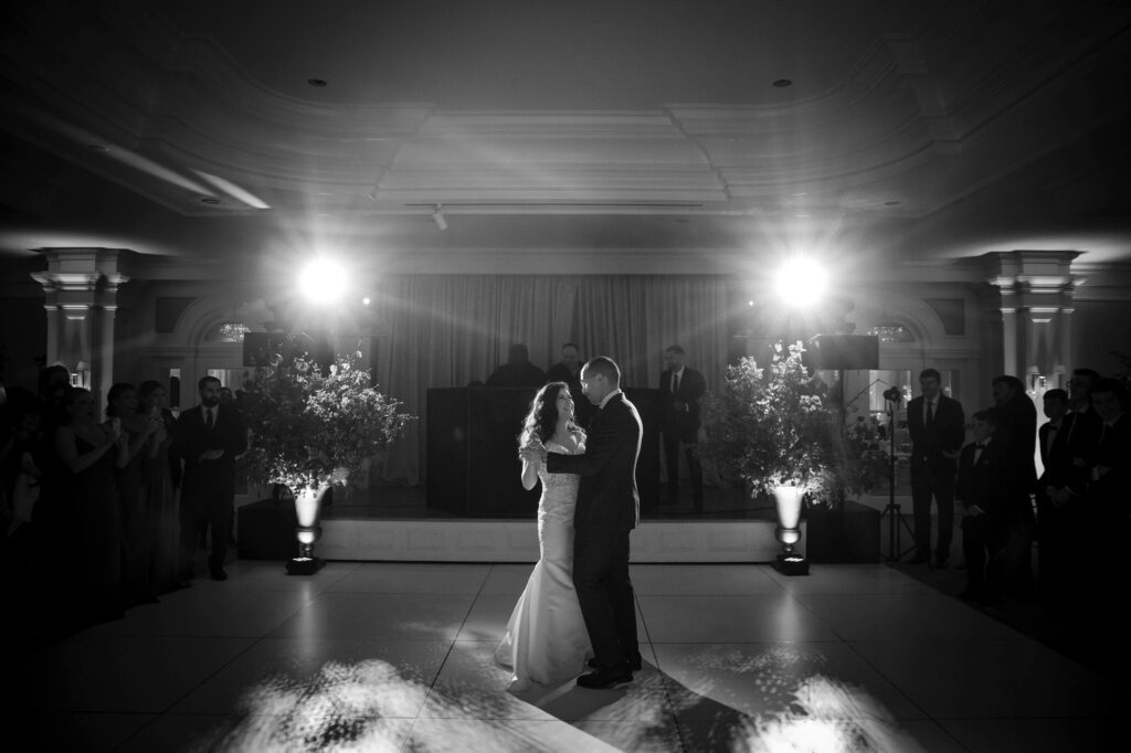 A couple shares their first dance at a Pine Hollow Country Club wedding in a dimly lit room with onlookers surrounding the dance floor.