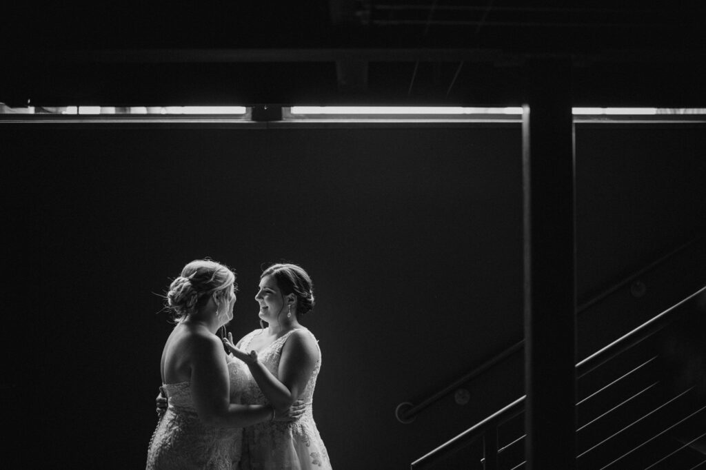 Two brides holding hands and smiling at each other, standing under a dark overpass with light illuminating them from the front, celebrating their saltwater Farms vineyard wedding.