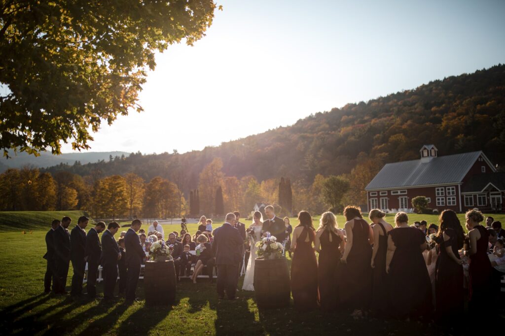 Outdoor wedding ceremony at Riverside Farm with guests and a couple exchanging vows, set against a backdrop of hills and a sunlit sky.