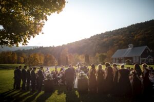 Outdoor wedding ceremony at Riverside Farm with guests and a couple exchanging vows, set against a backdrop of hills and a sunlit sky.