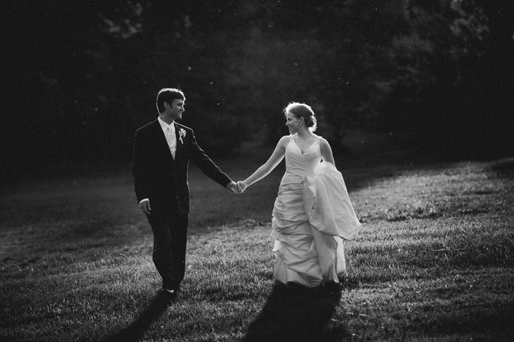 A black and white photo of a bride and groom holding hands and walking in a grassy field, warmly lit by sunlight during their Please Touch Museum wedding.