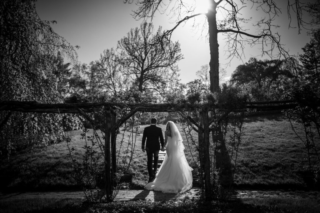 Newlyweds walking hand in hand under a pergola in a sunlit garden at Old Westbury Gardens.