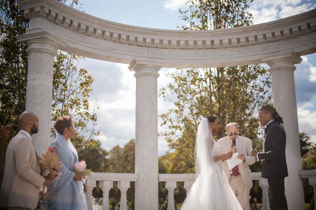 A wedding ceremony with a bride and groom exchanging vows under a white, classical arch at The Mansion on Main Street, surrounded by a small group of attendees on a sunny day.