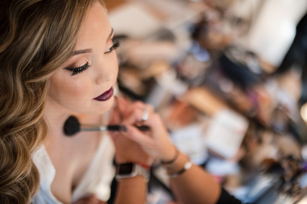 A woman having makeup applied by a brush, focusing on her lips, in preparation for a Rock Island Lake Club wedding, with a blurred salon background.