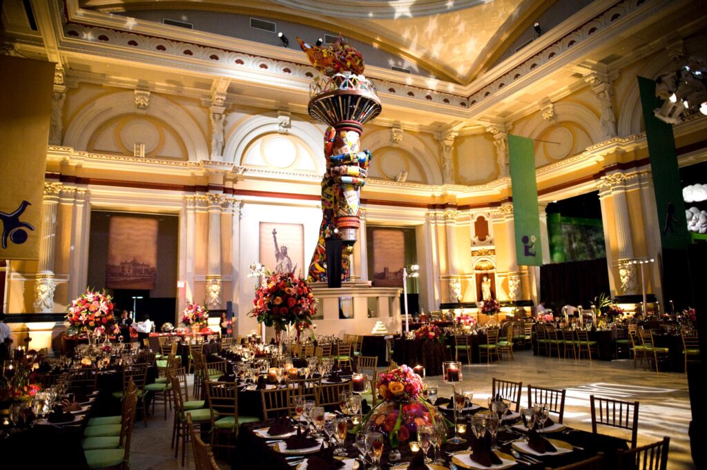 Elegant banquet setup in the grand hall of the Please Touch Museum with ornate ceilings, large floral arrangements, and a colorful central sculpture.