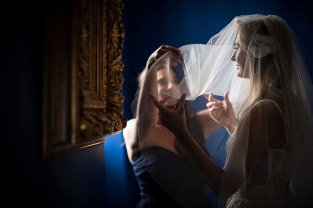 A bride in a white dress adjusts her veil with the help of a woman dressed in blue, reflected in an ornate mirror at a Congress Hall Cape May wedding, against a dark background.