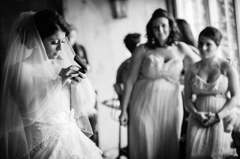 A bride in a white gown reads a heartfelt message on her phone at the Crossed Keys Inn in Andover, surrounded by bridesmaids in a softly lit room.