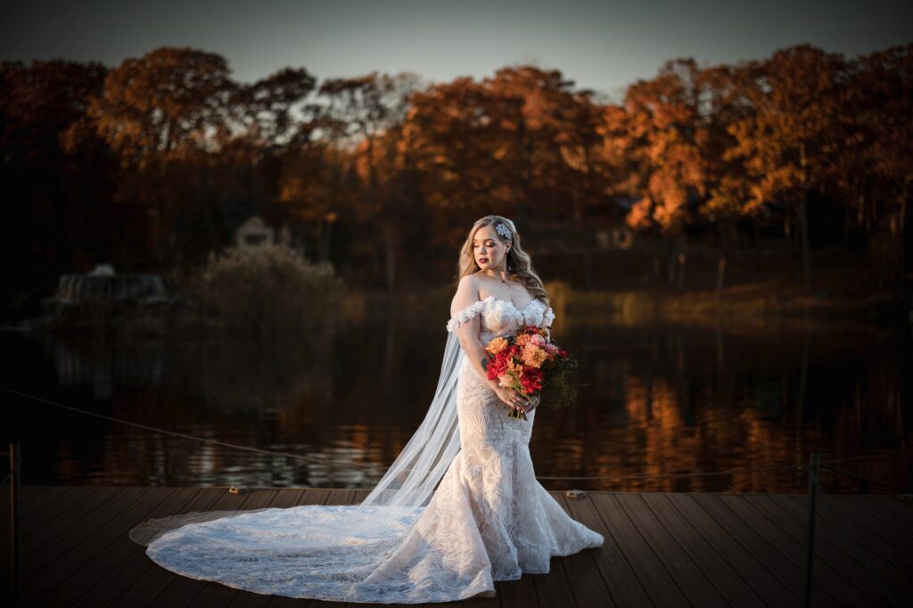 A bride in a lace gown stands on a dock by a lake at Rock Island Lake Club, holding a bouquet, with autumn trees in the background at sunset.