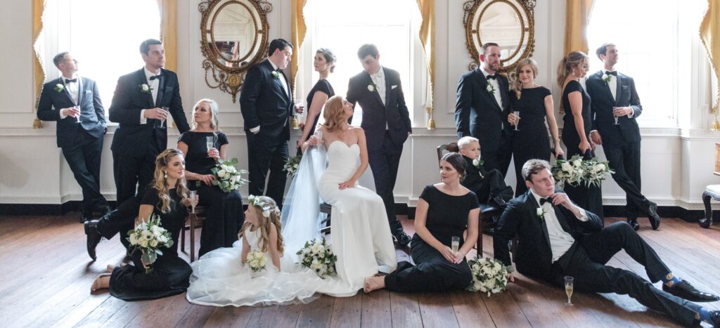 A wedding party posing elegantly in the Powel House's ornate room with lavish décor, chandeliers, and large mirrors, all dressed in formal black and white attire.