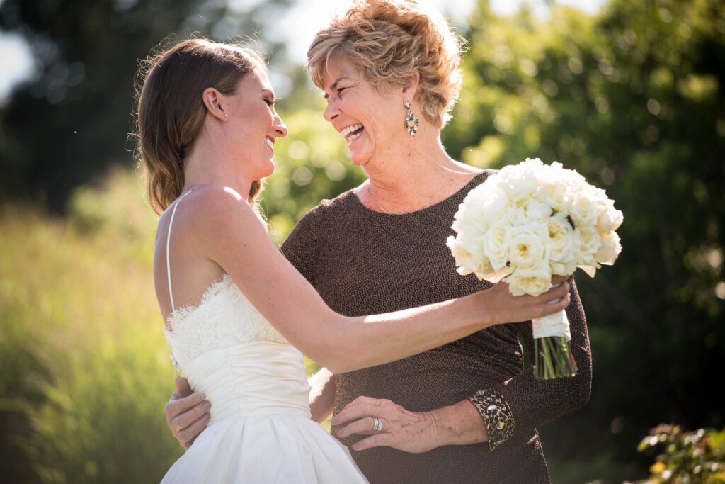 A bride sharing a joyful moment with her mom at an ocean house in Rhode Island, while holding a bouquet of white flowers.