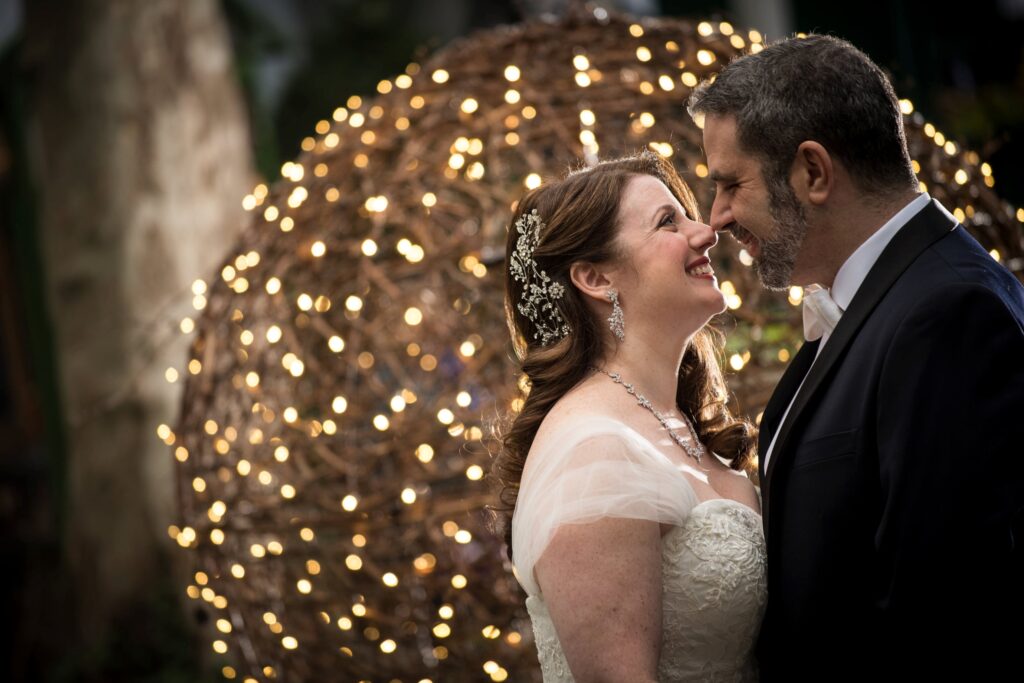 A bride and groom smiling at each other, standing in front of a large, illuminated orb made of fairy lights at their Bryant Park Grill NYC wedding.