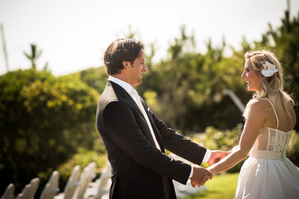 A bride and groom holding hands outdoors at Congress Hall Cape May, smiling at each other, with a green backdrop and sunlight.