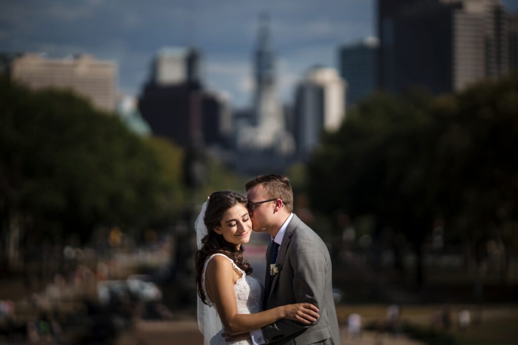 A bride and groom embracing and kissing at Water Works Philadelphia, with a city skyline in the background.