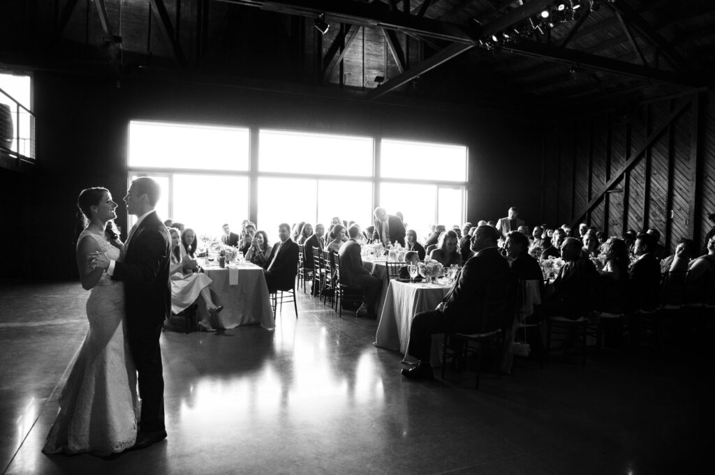 A bride and groom share a dance at their saltwater Farms vineyard wedding in a dimly lit hall, with guests seated at tables, backlit by large windows.