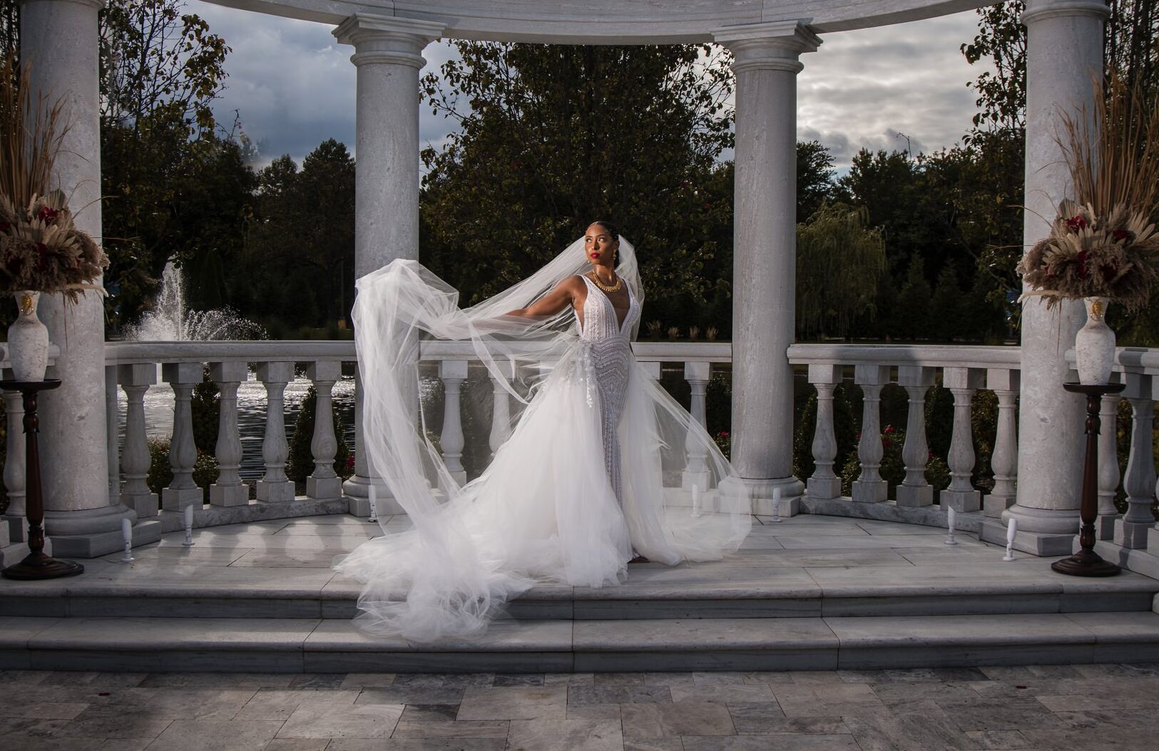 A bride stands under a neoclassical rotunda with columns at The Mansion on Main Street, her veil flowing, surrounded by decorative urns and trees against a cloudy sky.