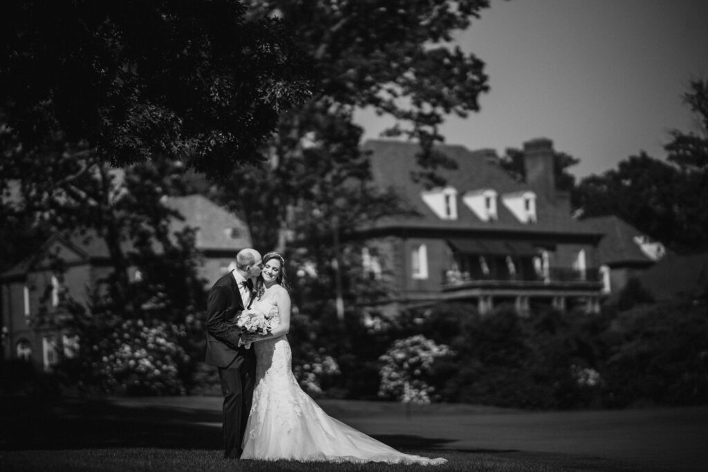 A bride and groom sharing a tender moment on a grassy lawn at the Pine Hollow Country Club, with a large house in the background.