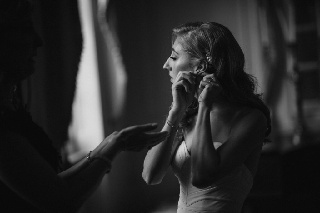 A monochrome image of a woman in a dress having her earring adjusted by another woman at a Powel House Philadelphia wedding, both illuminated by soft light.