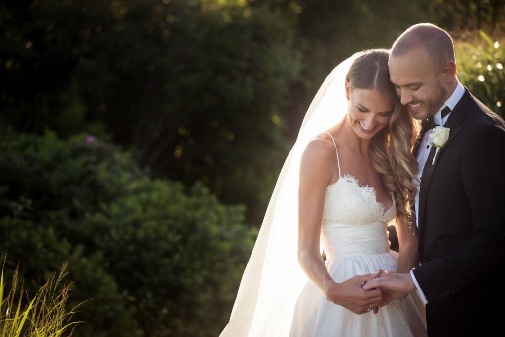 Bride and groom smiling and holding hands in an ocean house Rhode Island wedding, in a sunlit outdoor setting.