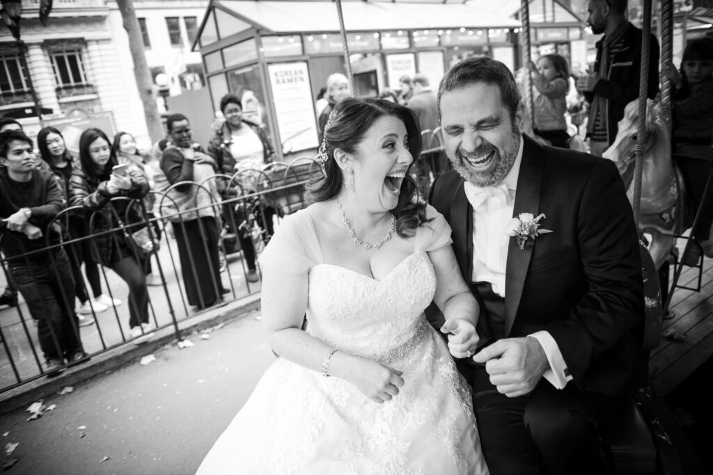 A joyful bride and groom laughing together on their wedding day at Bryant Park Grill NYC, surrounded by onlookers, in a black-and-white photo.