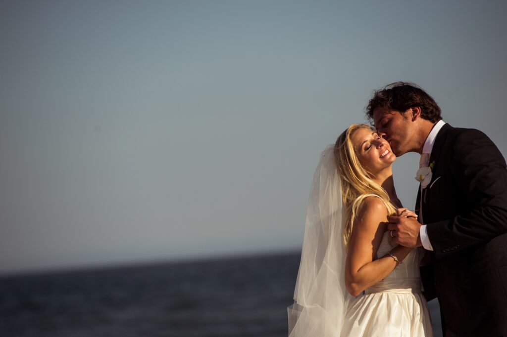 Bride and groom sharing a romantic kiss by the ocean under a clear sky at their Congress Hall Cape May wedding.
