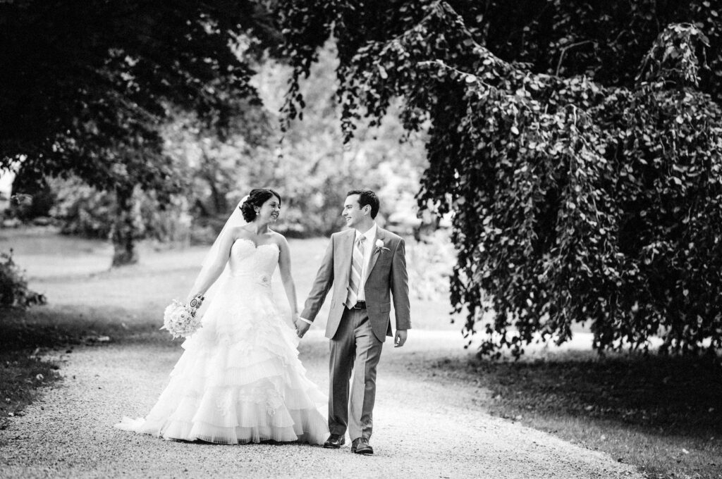 A bride in a ruffled gown and a groom in a grey suit holding hands and looking at each other in a tree-lined pathway at Crossed Keys Inn in Andover, photographed in black and white