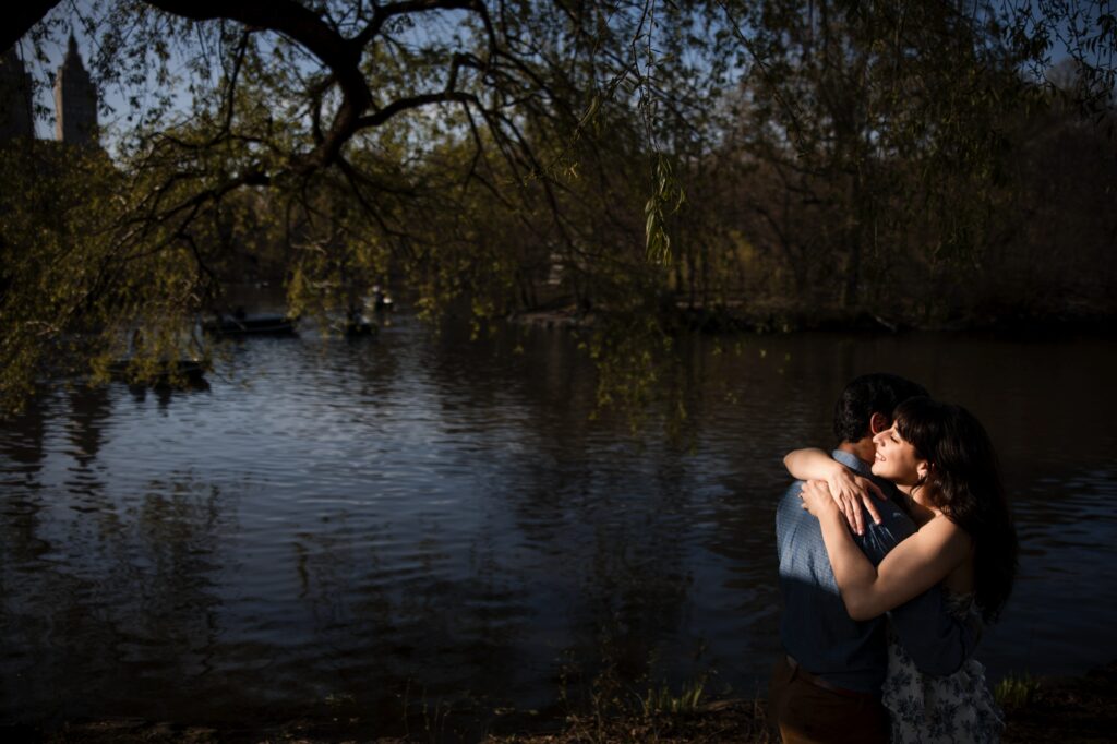 A couple embraces beside a lake under a tree in Central Park, with the sun filtered through branches and boats visible in the background.
