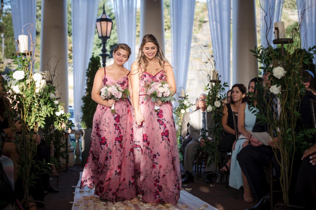 Two bridesmaids in floral dresses walking down the aisle at a Water Works Philadelphia outdoor wedding ceremony, holding bouquets.