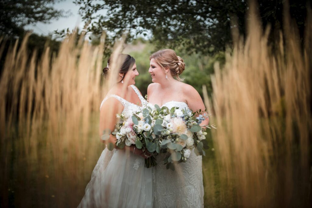 Two brides smiling at each other, holding a large floral bouquet, surrounded by tall grasses in a serene outdoor setting at Saltwater Farms Vineyard.