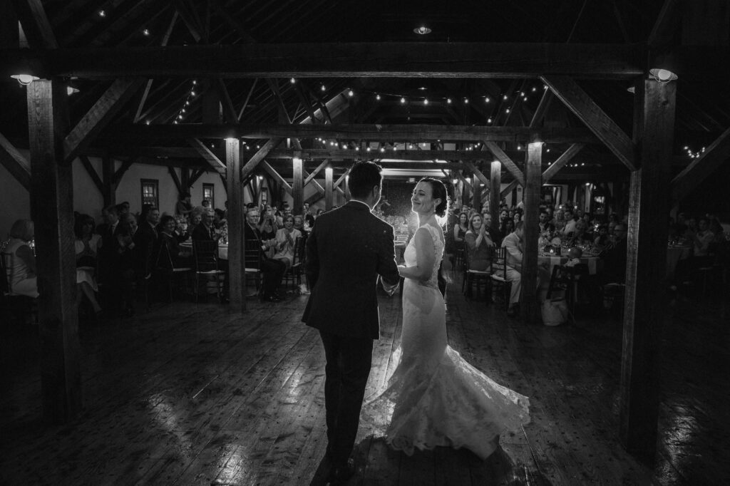 A bride and groom share their first dance at a Riverside Farm wedding in a rustic hall with wooden beams and onlookers seated around.