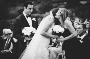A bride in a white gown hugs a woman holding flowers at her Resort at Pelican Hill wedding ceremony while the groom looks on, a crowd seated in the background.