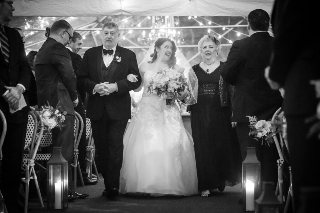 Bride and groom walking down the aisle at Bryant Park Grill NYC, smiling and holding hands, accompanied by an older couple, with guests and fairy lights in the background.