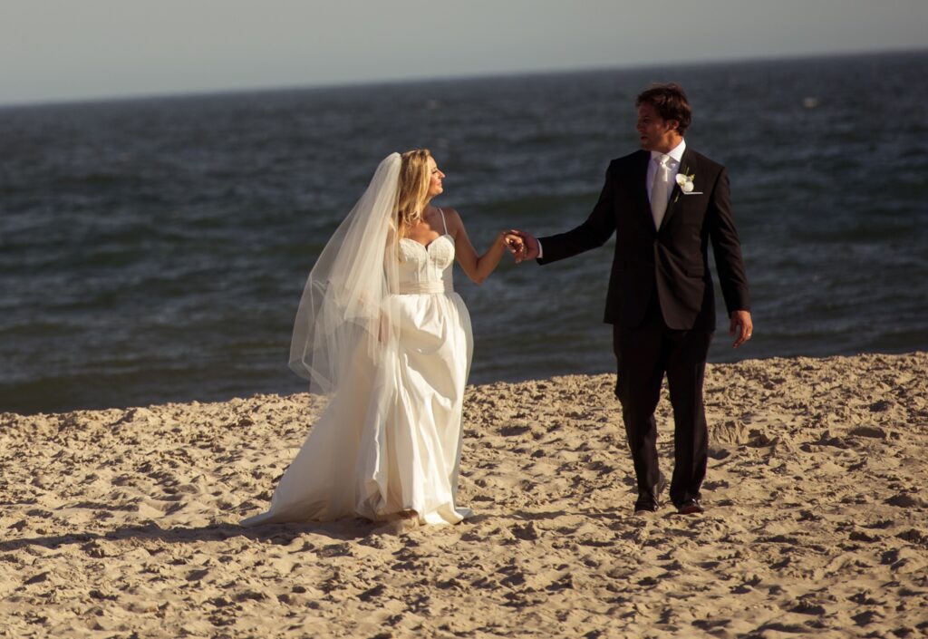 A bride and groom holding hands and walking along a sandy beach at Congress Hall Cape May, with the ocean in the background.