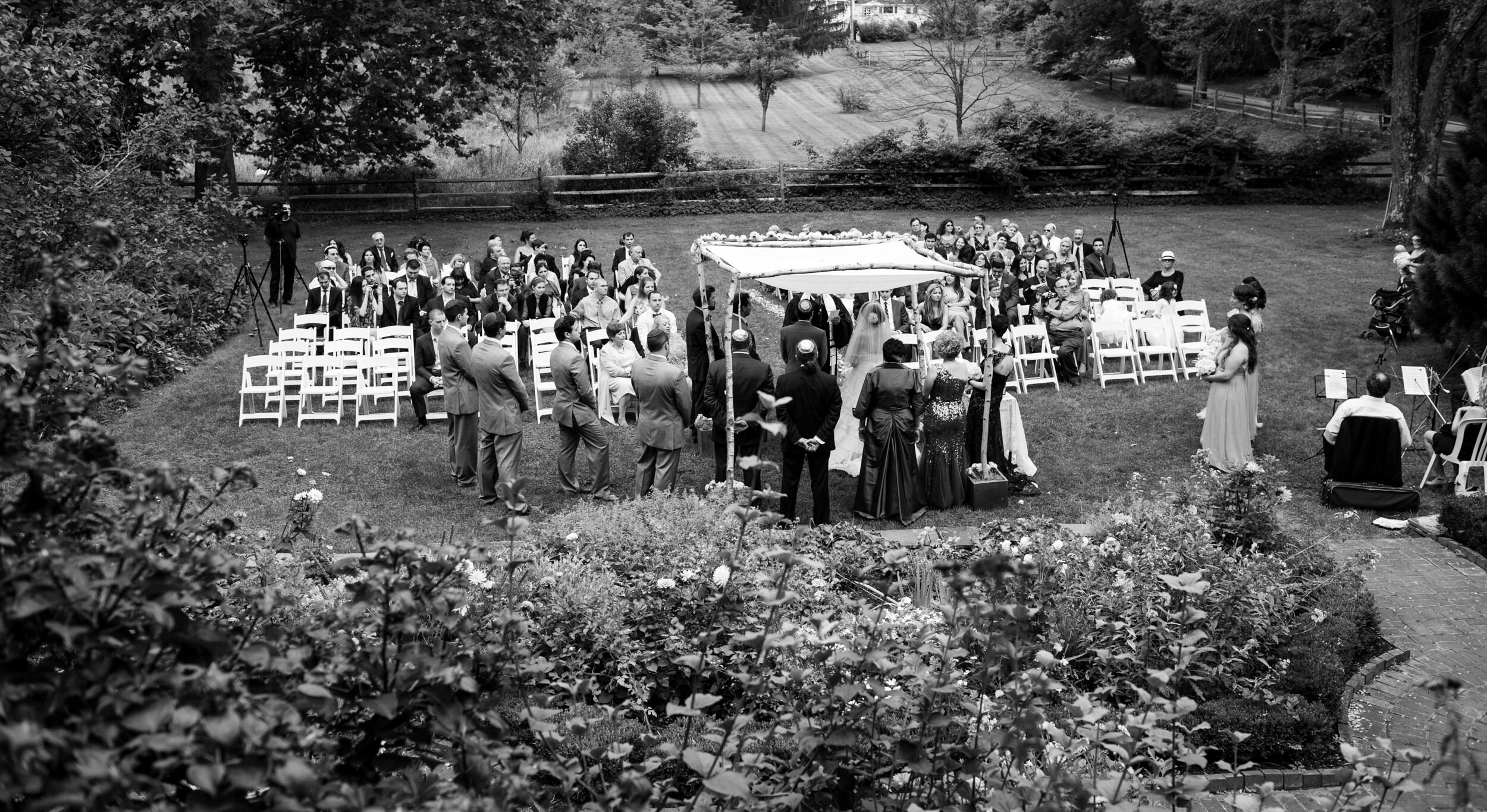 Black and white photo of an outdoor wedding ceremony at Crossed Keys Inn in Andover, with guests seated and standing around a couple under a floral arch.