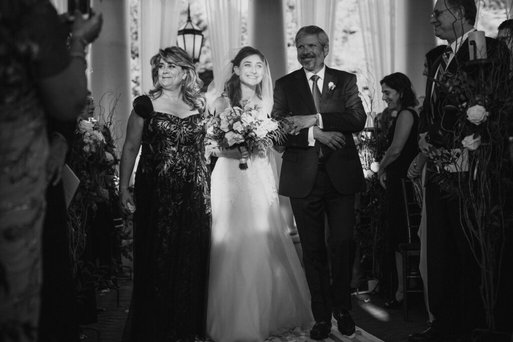 A bride walks down the aisle with her parents on either side, all smiling happily in a black-and-white photo at a Water Works Philadelphia wedding.