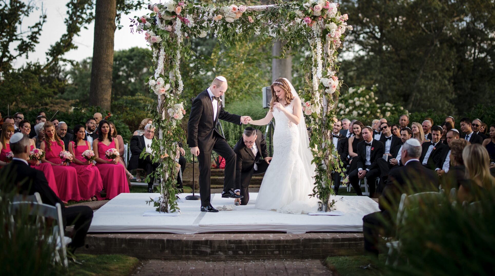 A couple holds hands at the altar during an outdoor wedding ceremony at Pine Hollow Country Club as guests look on.