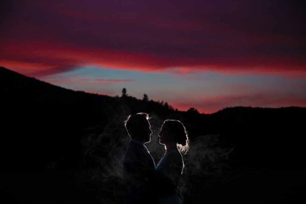 Silhouetted couple with a vibrant sunset and mountains in the background at a Riverside Farm wedding, highlighting a romantic scene.