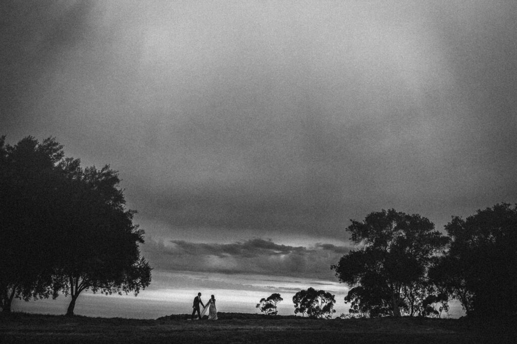 Two people walking between trees under a cloudy sky at the Resort at Pelican Hill wedding, with beams of light shining through the clouds.
