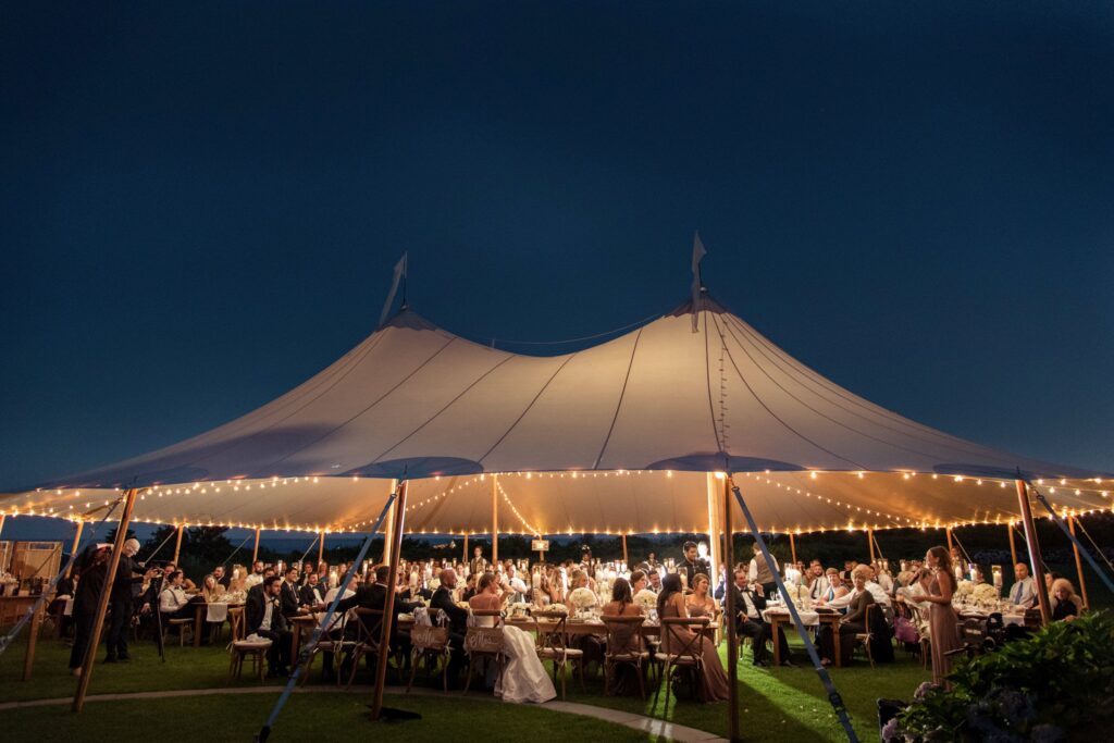 An open-air evening event at Ocean House Rhode Island under a large tent adorned with string lights, with guests seated at tables for a wedding.