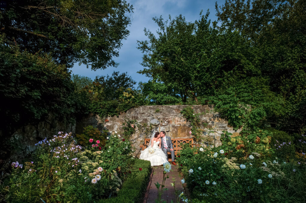 A newlywed couple sharing a kiss on a quaint garden path at Crossed Keys Inn Andover, surrounded by lush flowers and greenery under a clear sky.