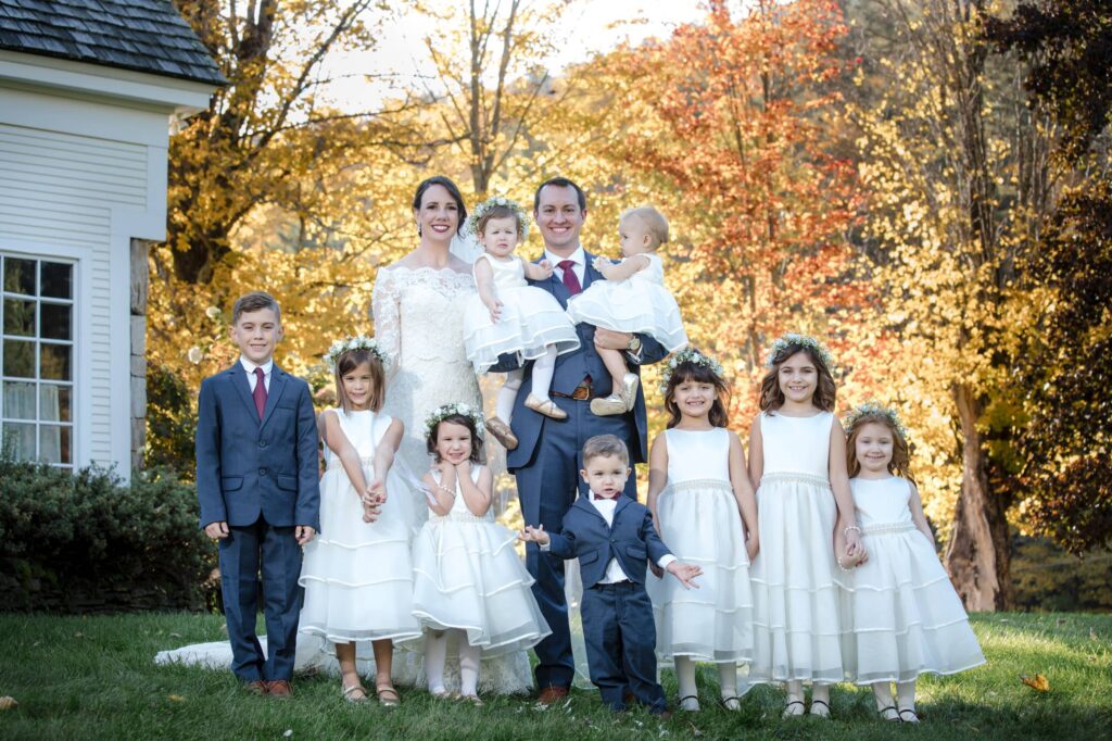 A family portrait outdoors with two adults and seven children, wearing formal attire at a Riverside Farm wedding, standing and sitting in front of trees with autumn foliage.